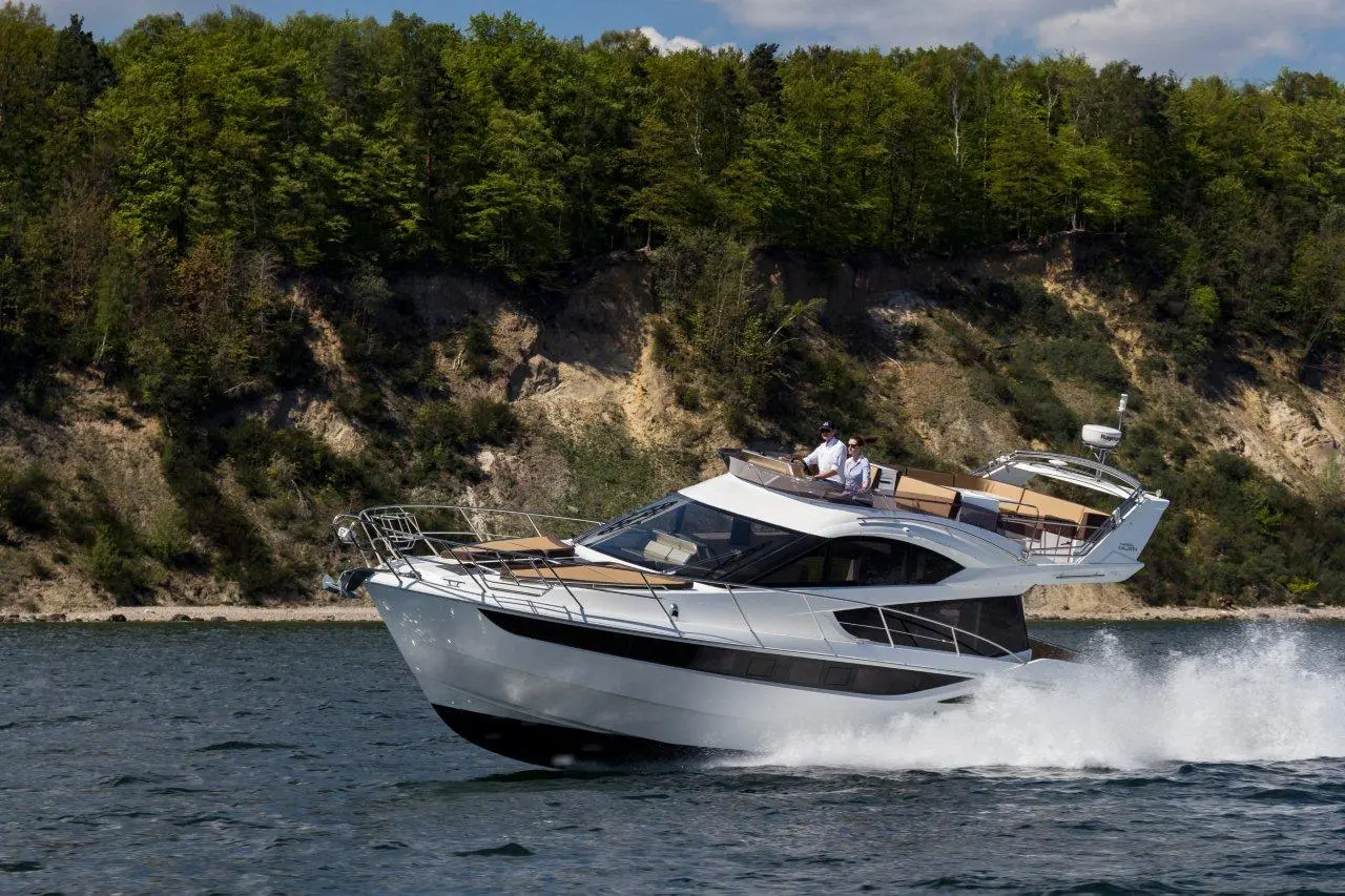 A sleek white motor yacht speeds over water, with two people at the helm and lush, green trees in the background.