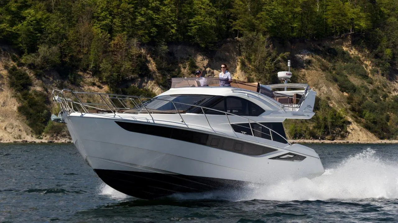 A sleek motor yacht speeds through calm waters, with two people enjoying the view from the bridge under lush greenery.