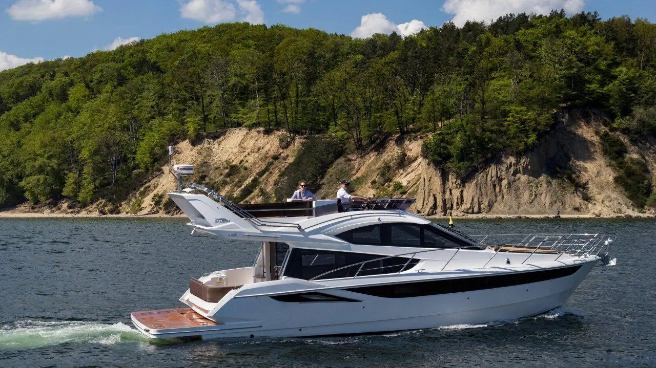 A sleek white yacht cruises on calm waters near a lush green shoreline, with two men at the helm enjoying the scenery.