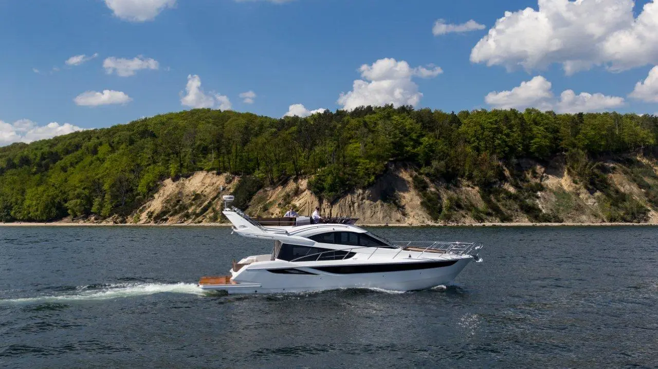 A luxury motor yacht glides on calm water near a green hillside, with two people enjoying the view on the upper deck.