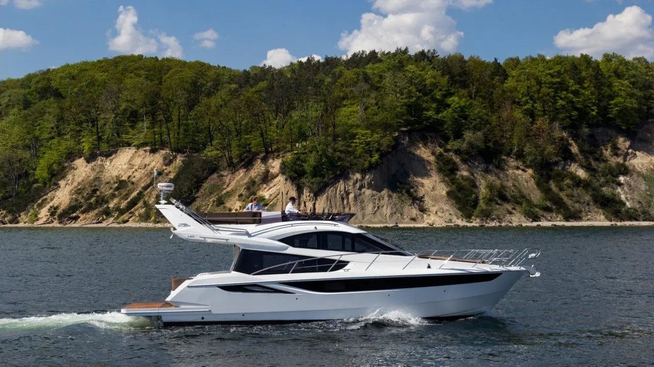 A sleek white yacht navigates a serene body of water, with lush green hills and a sandy shoreline in the background.