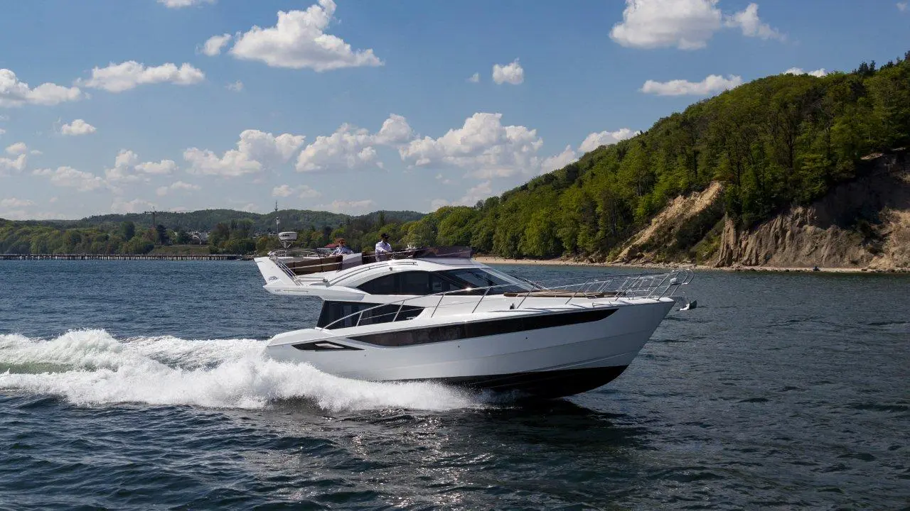 A sleek motor yacht cruising on a calm waterway with green hills and a blue sky in the background.