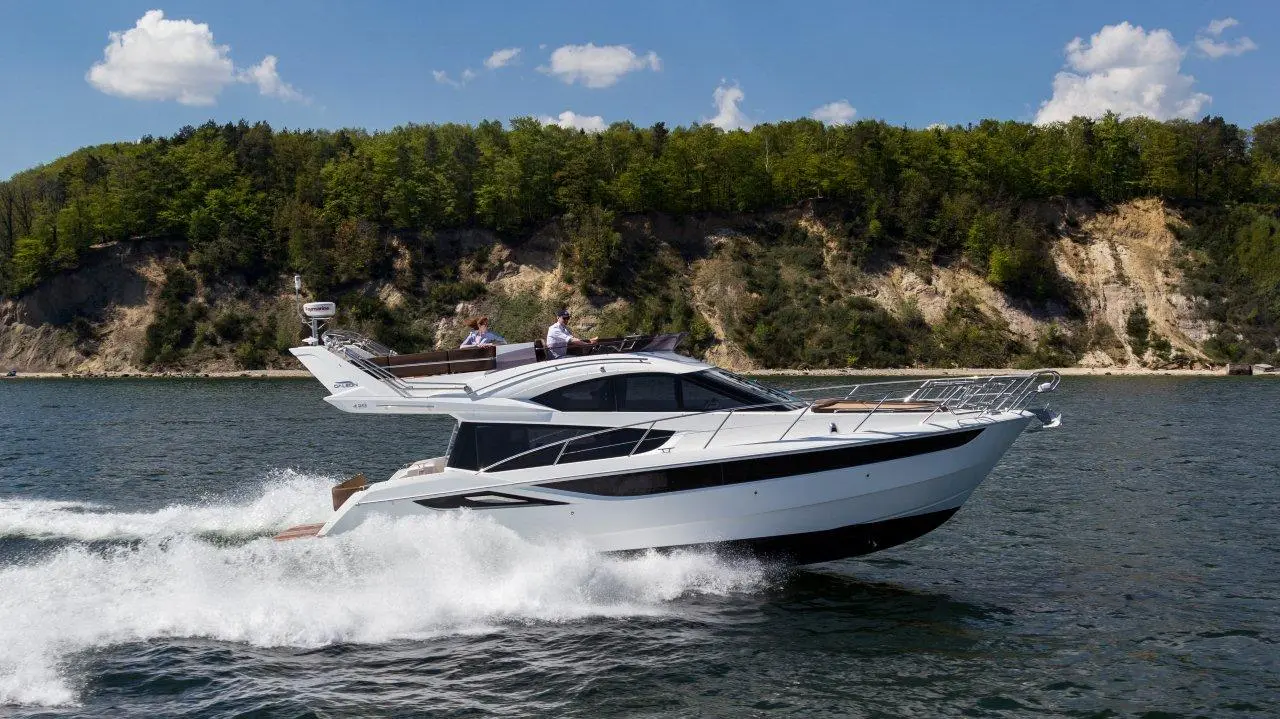 A sleek white motorboat speeds through water, surrounded by greenery and cliffs under a clear blue sky.