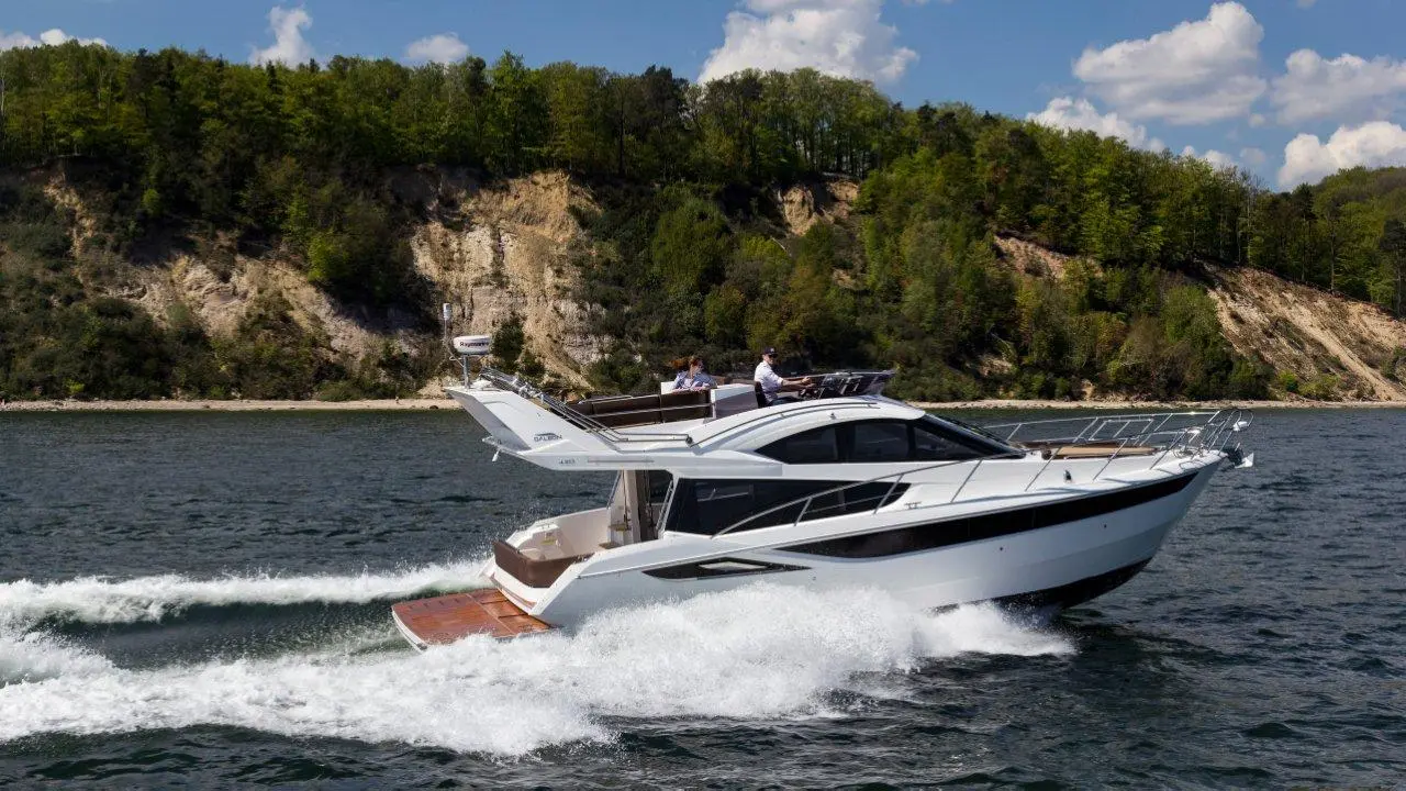 A sleek motorboat cruising on a river, with two people on board, surrounded by green hills and a clear blue sky.
