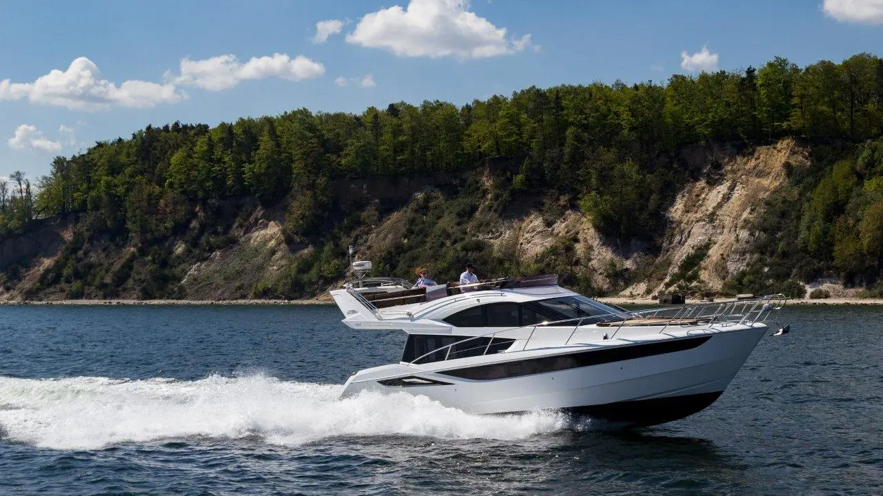 A white luxury yacht speeds across a serene lake, surrounded by lush green trees and steep cliffs under a blue sky.