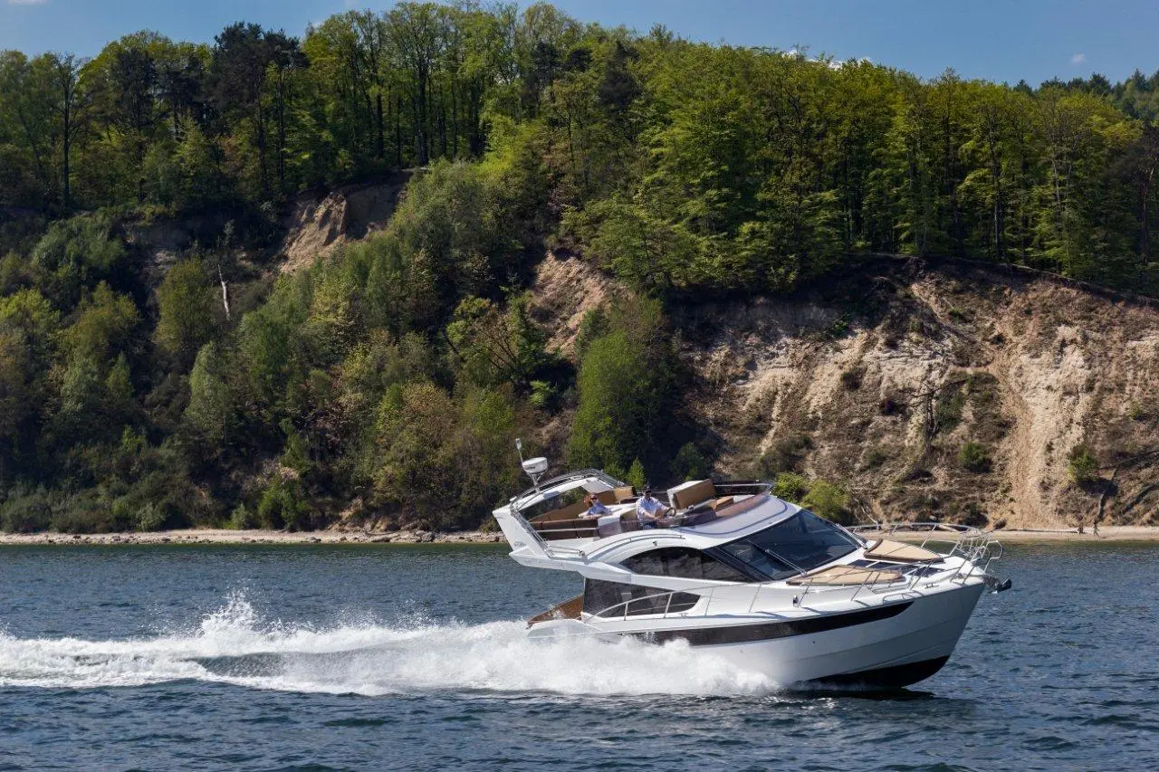 A white luxury motorboat glides across calm waters with a lush green forest in the background, showcasing a scenic landscape.