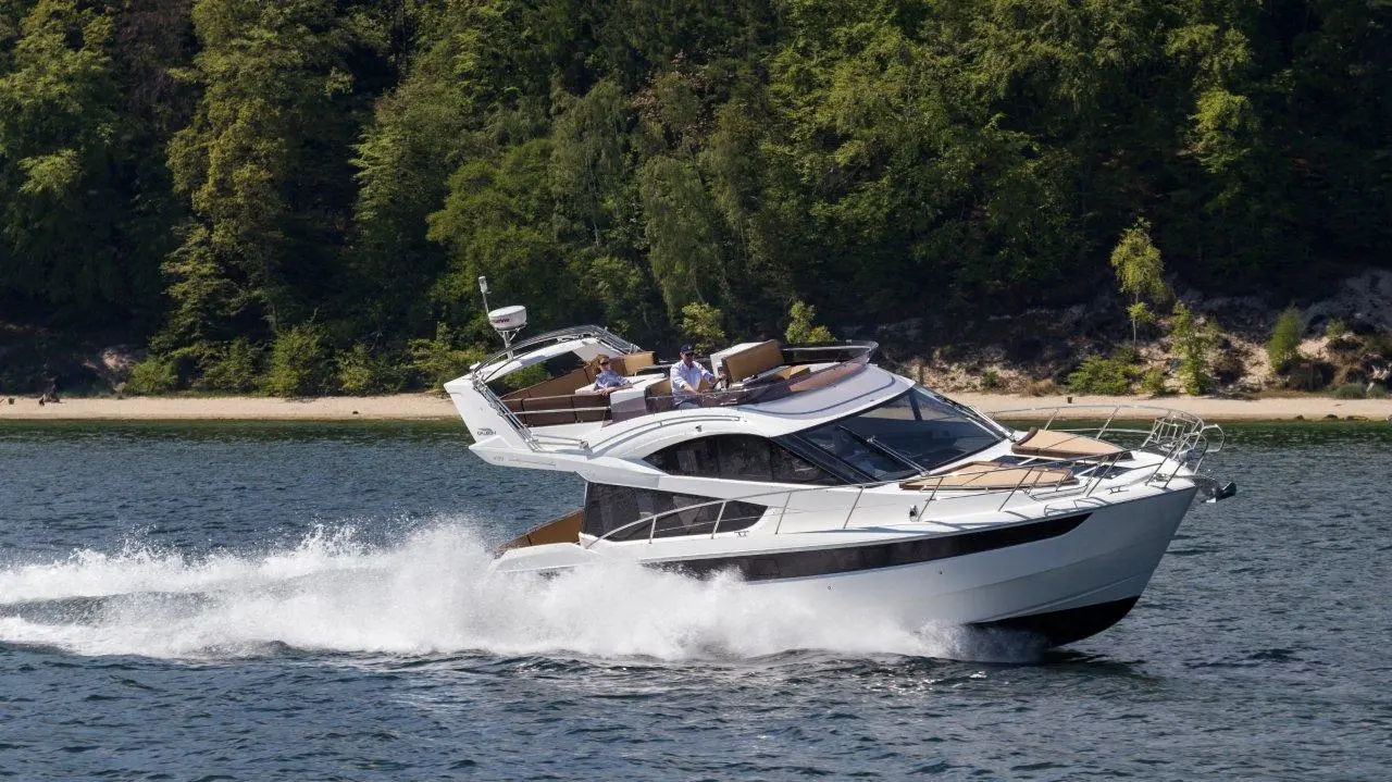 A sleek white yacht speeds across a calm lake, with two people enjoying the ride, surrounded by lush green trees and a sandy shore.