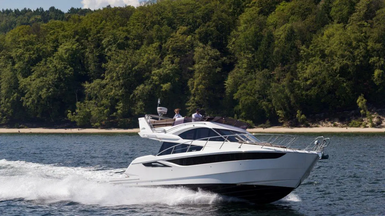 A sleek white motorboat speeds across the water, with two people on board and a lush green shoreline in the background.