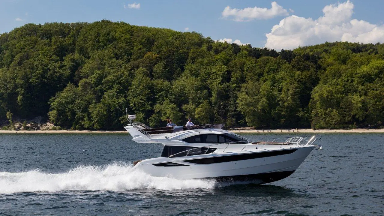A sleek white motor yacht navigates through calm waters, with green wooded shores and a sandy beach in the background.