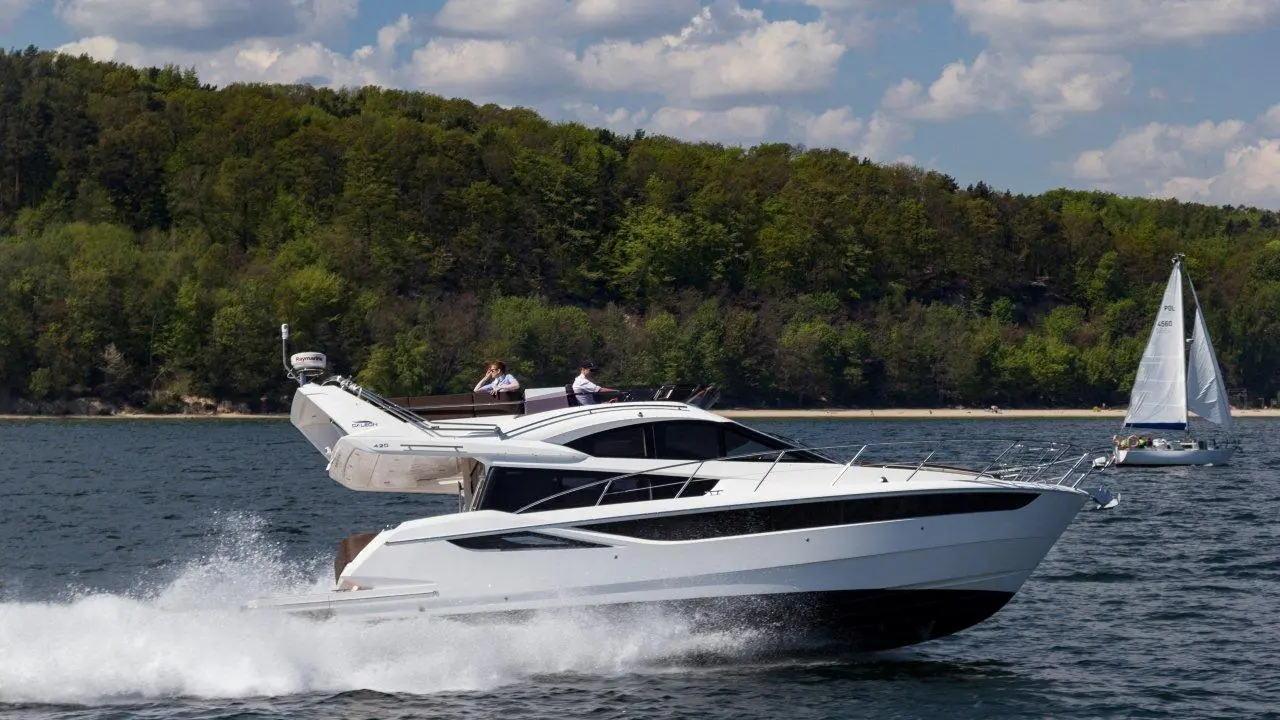 A white motor yacht speeds across the water, with lush green hills in the background and a sailboat nearby.