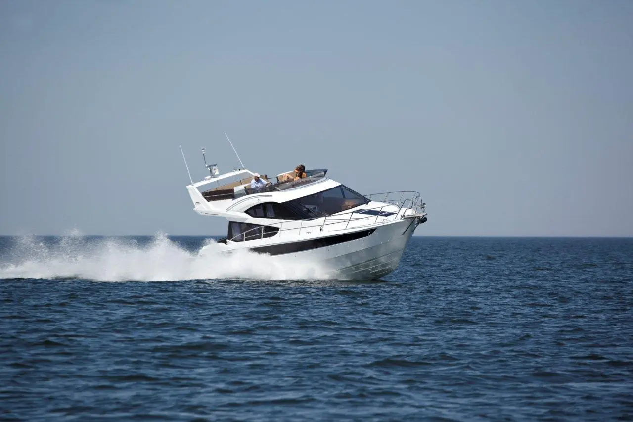 A white motor yacht speeds across calm blue waters, with two people visible on the deck enjoying the ride.