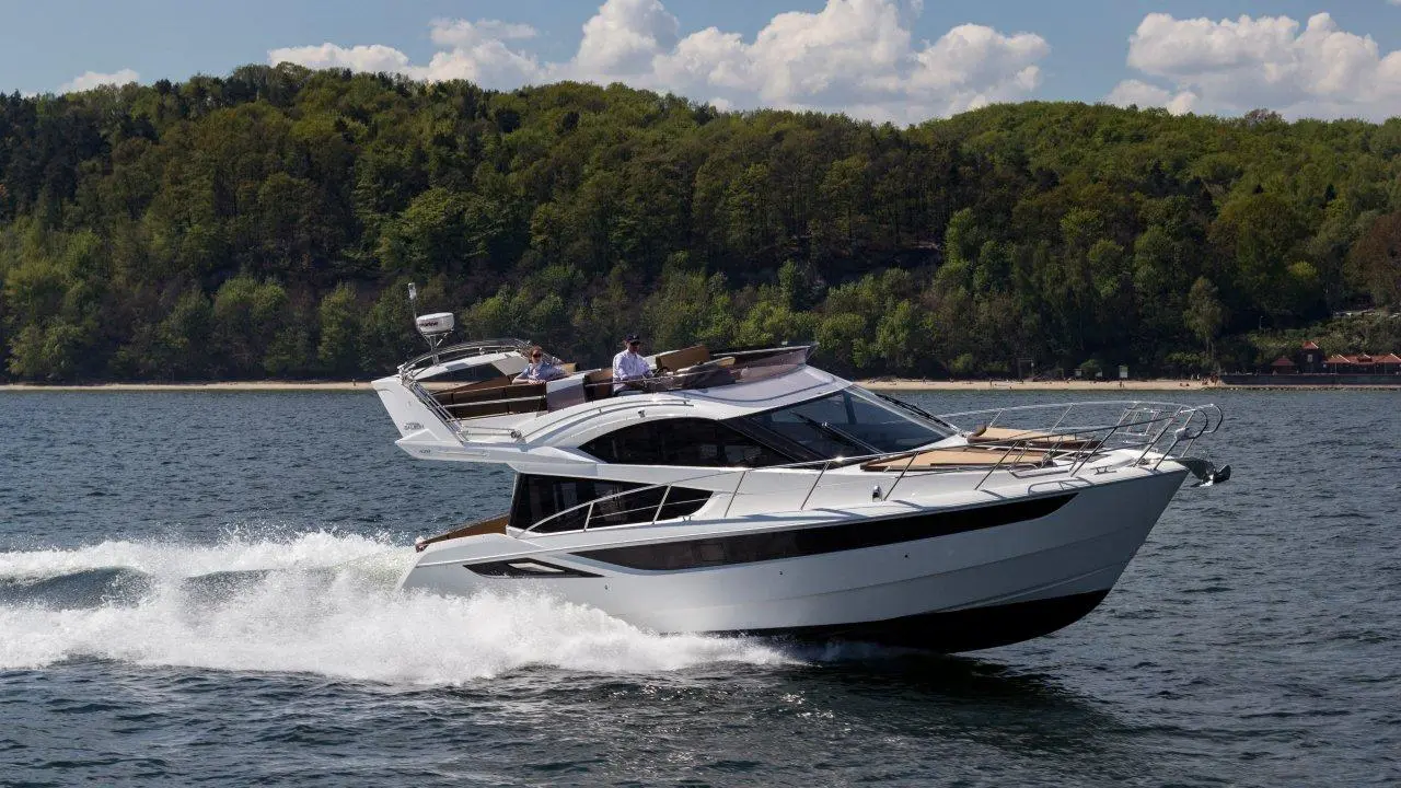 A luxury motor yacht speeds across a blue lake, with green hills and a clear sky in the background. Two men are onboard.