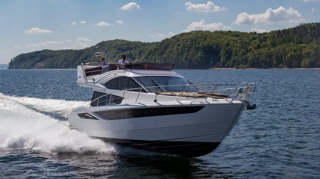 A luxury motor yacht speeds across a calm lake under blue skies, with two people at the helm enjoying the ride.