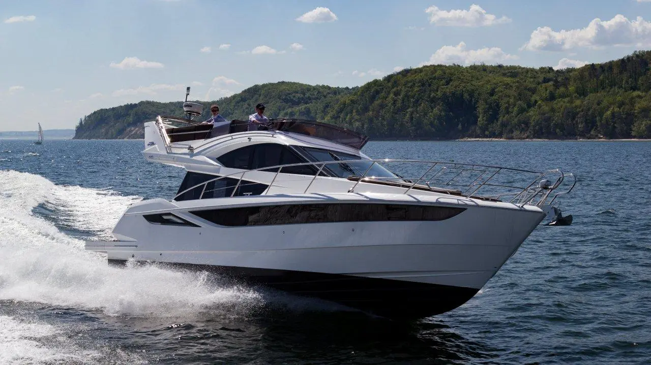 A sleek motor yacht navigates through water with two men at the helm, surrounded by green hills and blue skies.
