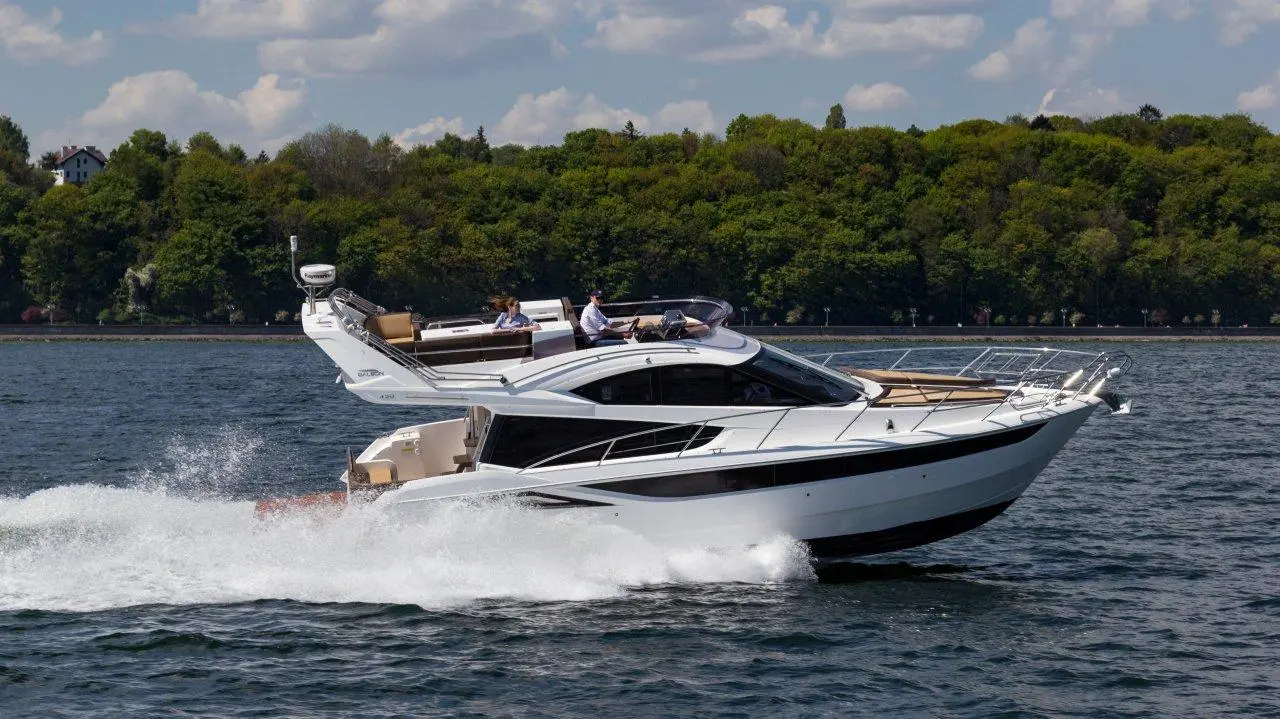 A stylish motorboat navigates through water, creating waves, with people enjoying the sunny day and greenery in the background.