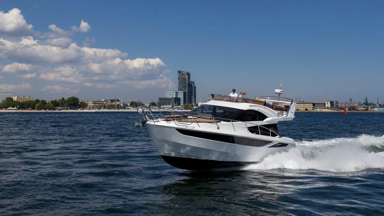 A sleek motor yacht speeds through waves, with a city skyline and clouds in the background, suggesting a sunny coastal day.