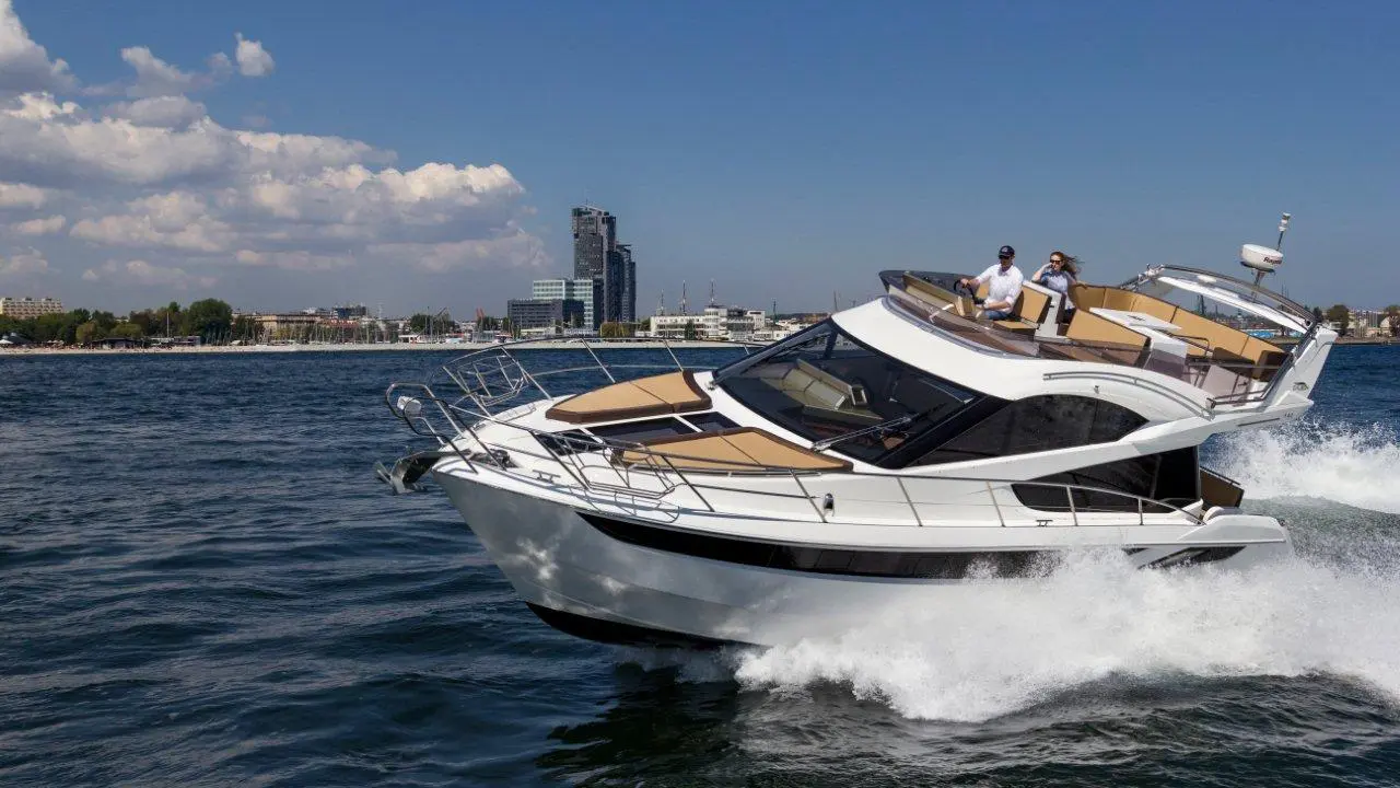 A white motor yacht speeds through blue water, with two people at the helm and a city skyline in the background.