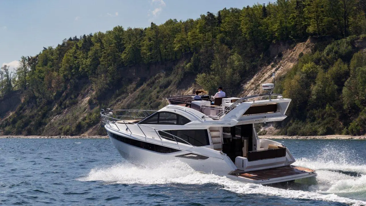 A sleek motor yacht gliding through clear blue waters, surrounded by lush green hills and a sunny sky.