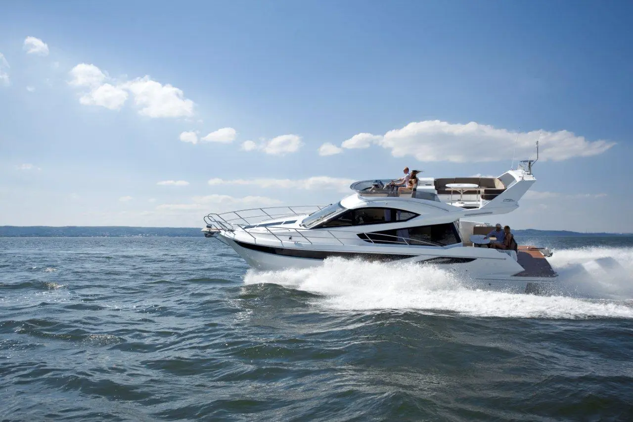 A sleek white yacht speeds through blue waters under a clear sky, with people enjoying the sun on board.