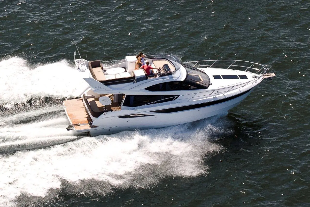 A sleek white luxury motor yacht cruises over shimmering blue waters, with two passengers enjoying the ride.