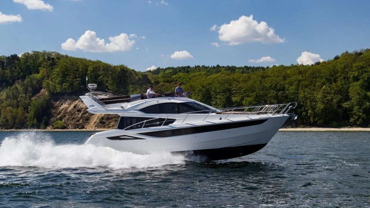A sleek white yacht speeds across calm water, with two people enjoying the sun on its deck and lush green hills in the background.