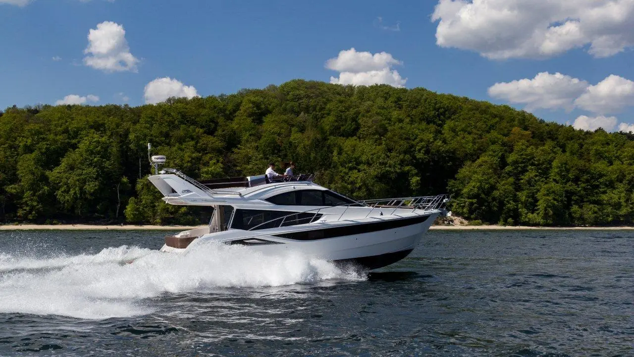 A sleek white motorboat speeds across the water, with two people at the helm and a forested shoreline in the background.