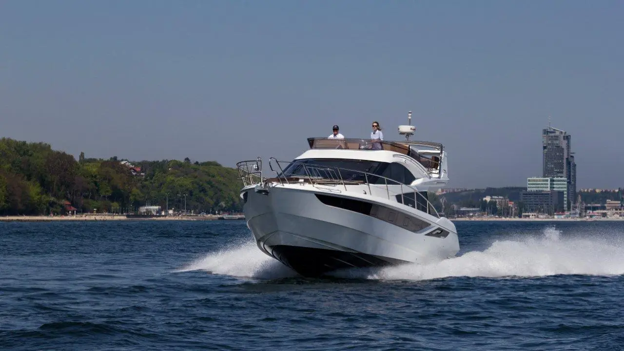A sleek white motor yacht glides over the water, with two people on deck and a city skyline in the background.