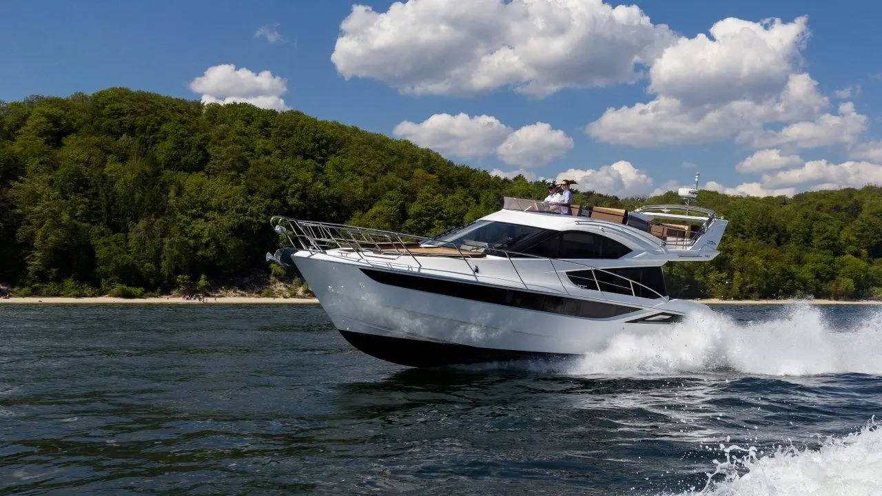 A sleek white motor yacht cuts through water, creating a wake, with a lush green forested shoreline and fluffy clouds above.