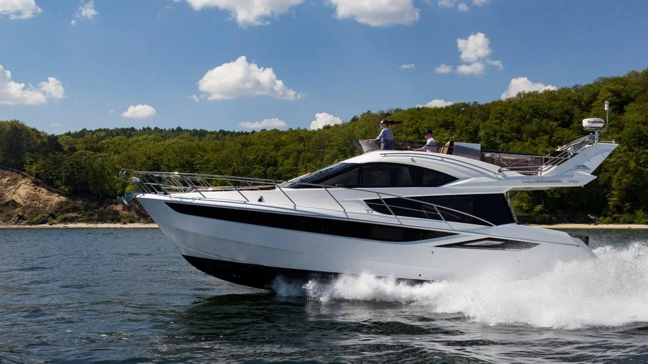 A sleek white yacht gliding through calm waters, with two people on board enjoying a sunny day. Lush greenery in the background.