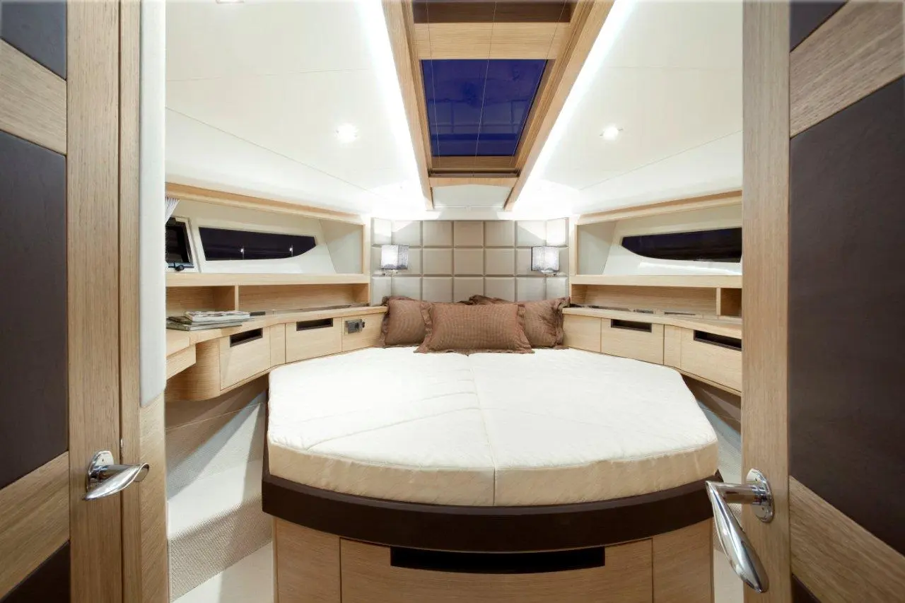 Cozy yacht cabin featuring a circular bed, wooden furniture, and soft lighting with shelves and a skylight above.