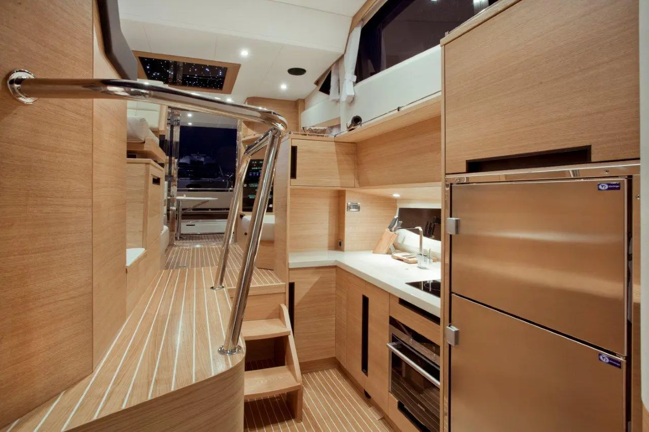 Interior view of a modern yacht galley with wooden cabinetry, a stainless steel fridge, and a view through a large window.