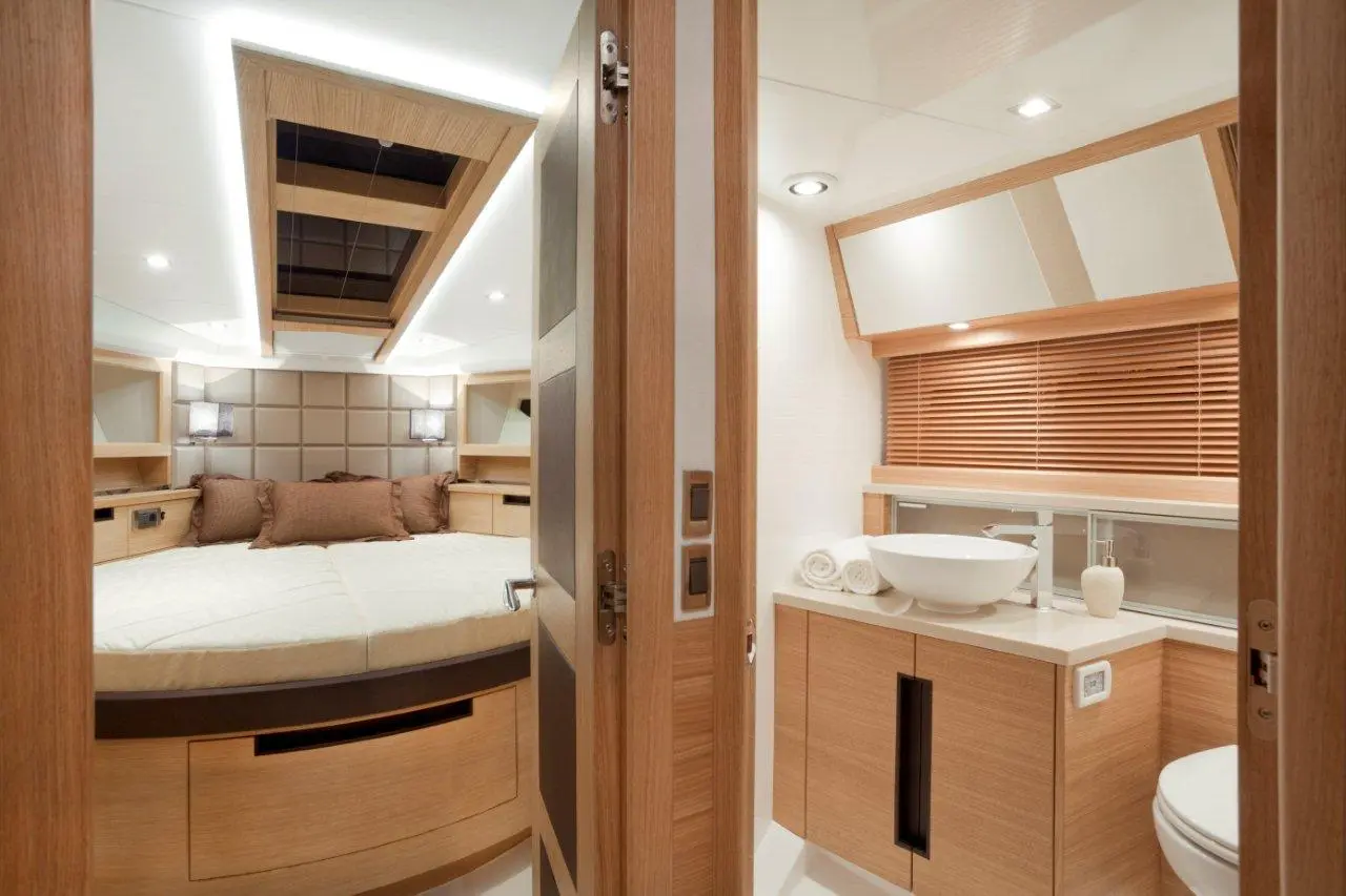 Luxurious boat cabin featuring a round bed, wooden storage, and a modern bathroom with a sink and towels. Bright lighting throughout.