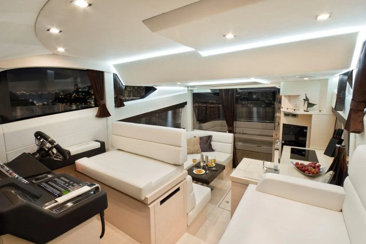 Luxury yacht interior featuring white seating, a control panel, and a dining table with food and drinks, illuminated by soft lighting.