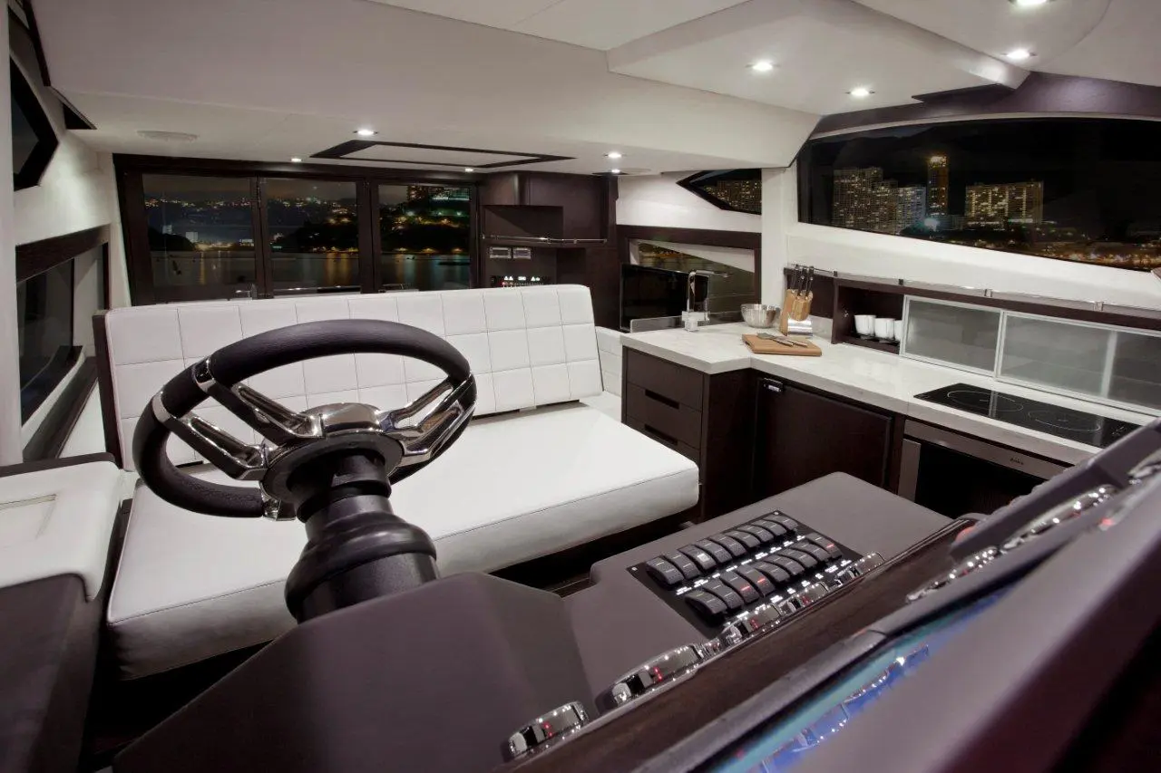 Modern yacht interior featuring a steering wheel, sleek white seating, and a stylish kitchen with city views at night.