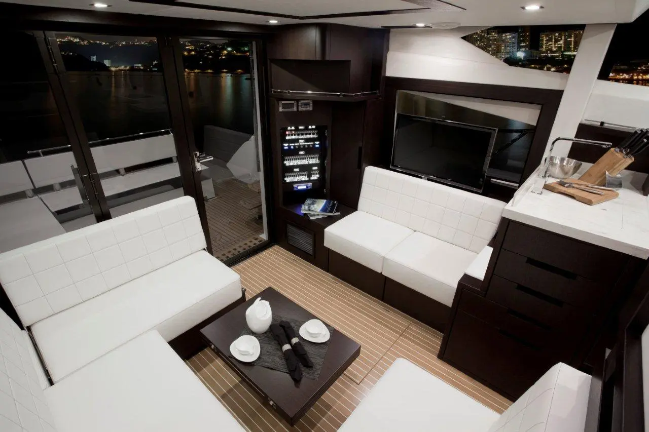 Luxurious yacht interior with white seating, wooden floor, a modern kitchen area, and a view of the city at night.