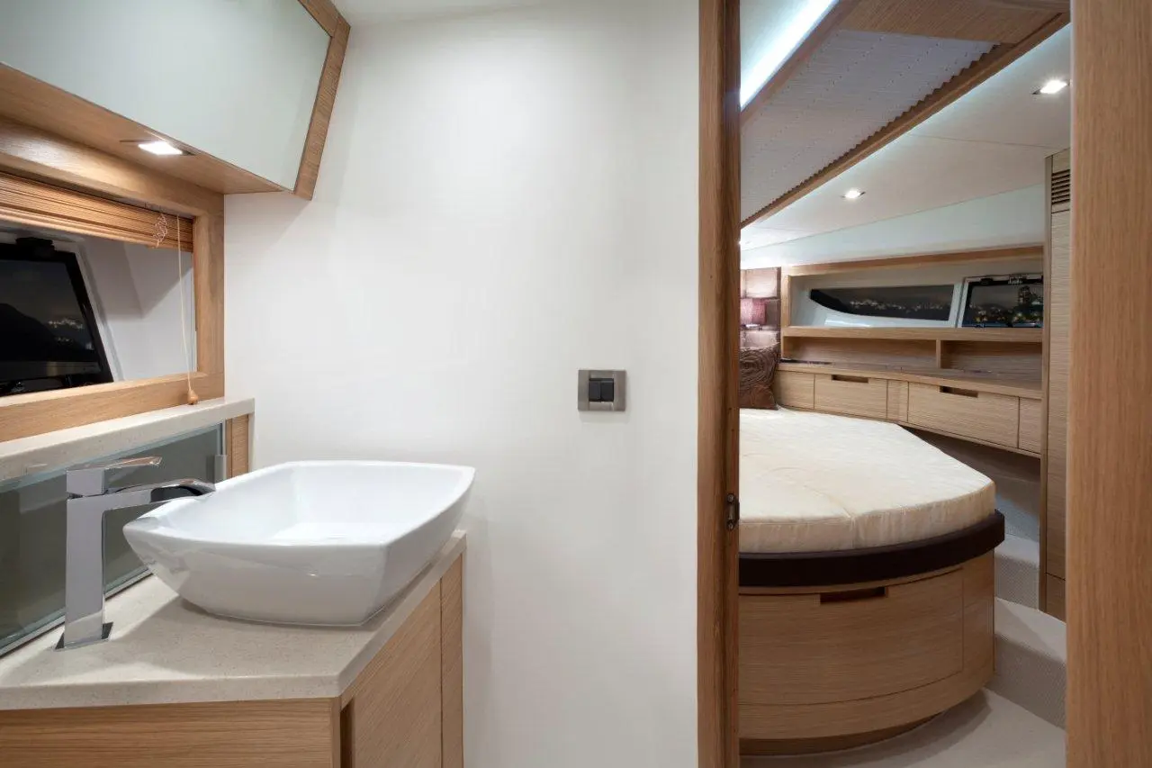 Modern yacht interior showing a sleek bathroom with a white basin, wooden cabinetry, and a cozy sleeping area visible beyond.