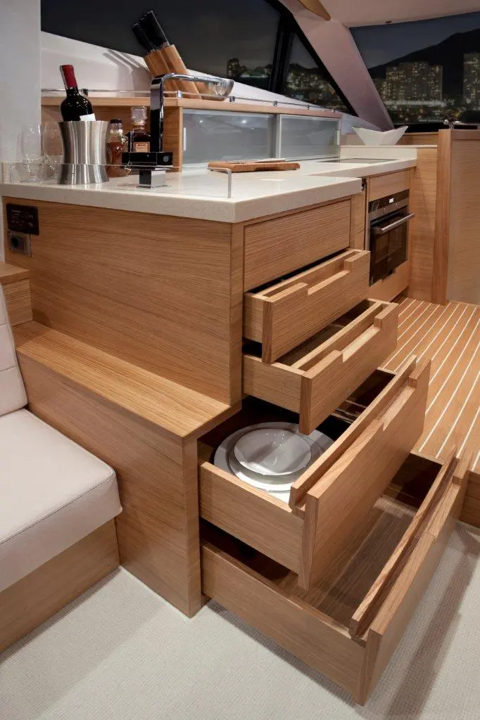 Modern wooden kitchen unit with open drawers displaying plates, wine bottles and a kitchen tool holder, set in a boat interior.