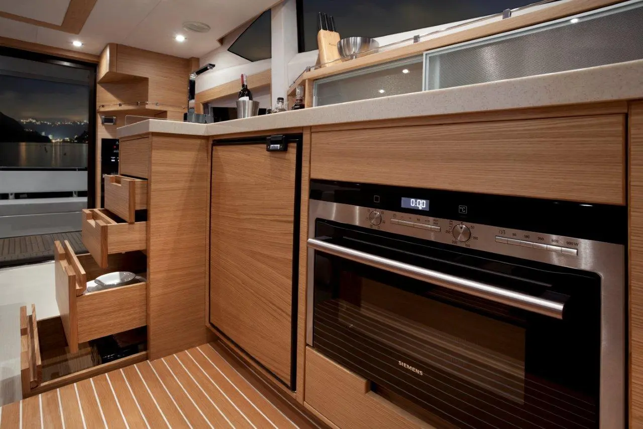 Modern yacht kitchen with wooden cabinetry, an oven, open drawers, and a view of the night sea through the window.