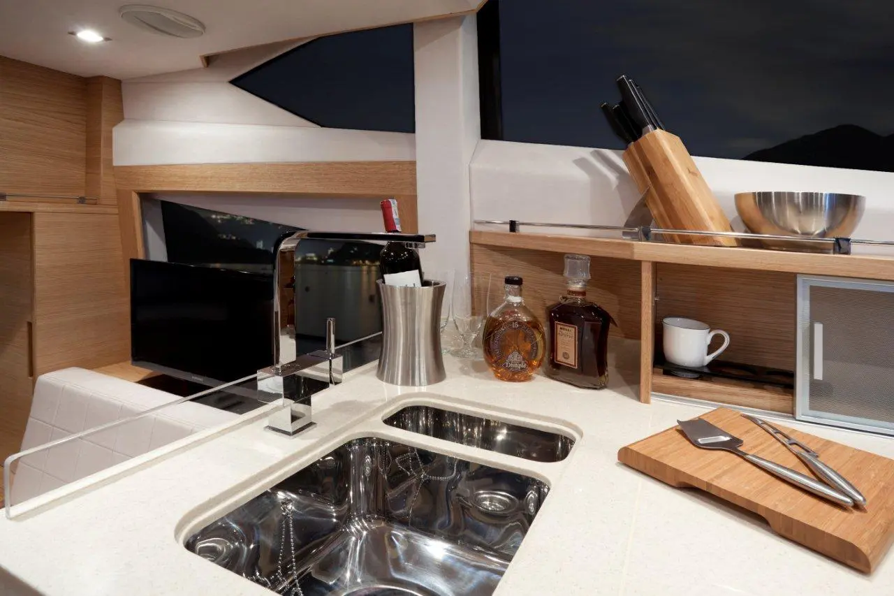 Modern yacht kitchen with stainless sink, wine bottles, glassware, and a chopping board, illuminated by soft lighting.
