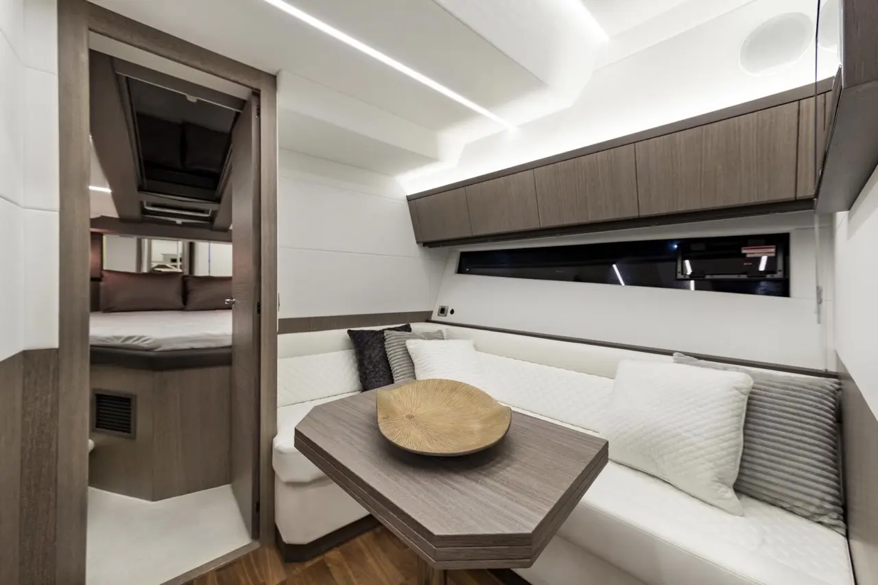 Modern yacht interior featuring a seating area with a wooden table, decorative pillows, and a doorway leading to a cozy bedroom.