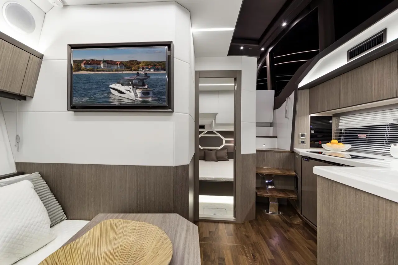 Modern yacht interior featuring a cozy seating area, kitchen, and a TV displaying a boat on water. Natural wood finishes throughout.