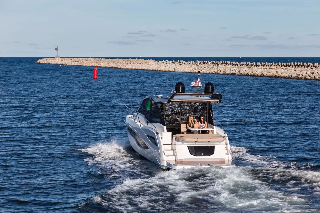A luxury boat with two women on board cruising through water near a breakwater and navigational buoys in a clear sky.