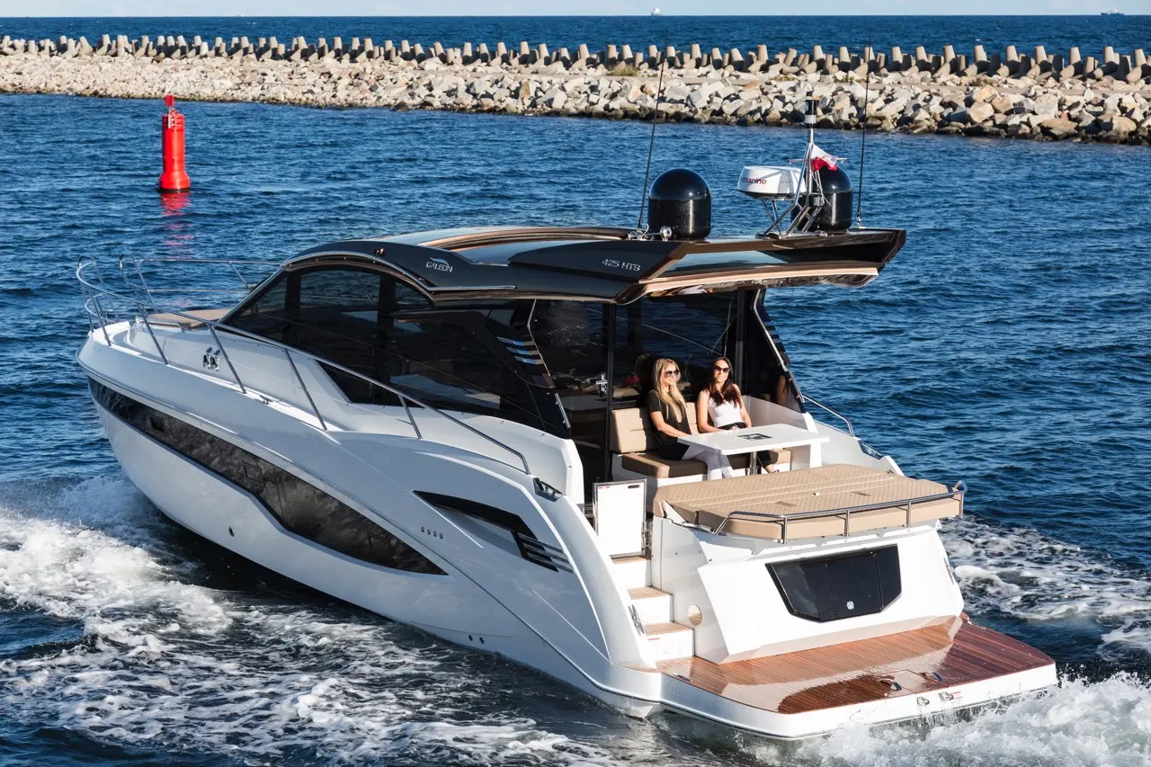 A luxury boat with two women sitting at a dining table, cruising on blue water near a rocky shoreline with a red buoy.