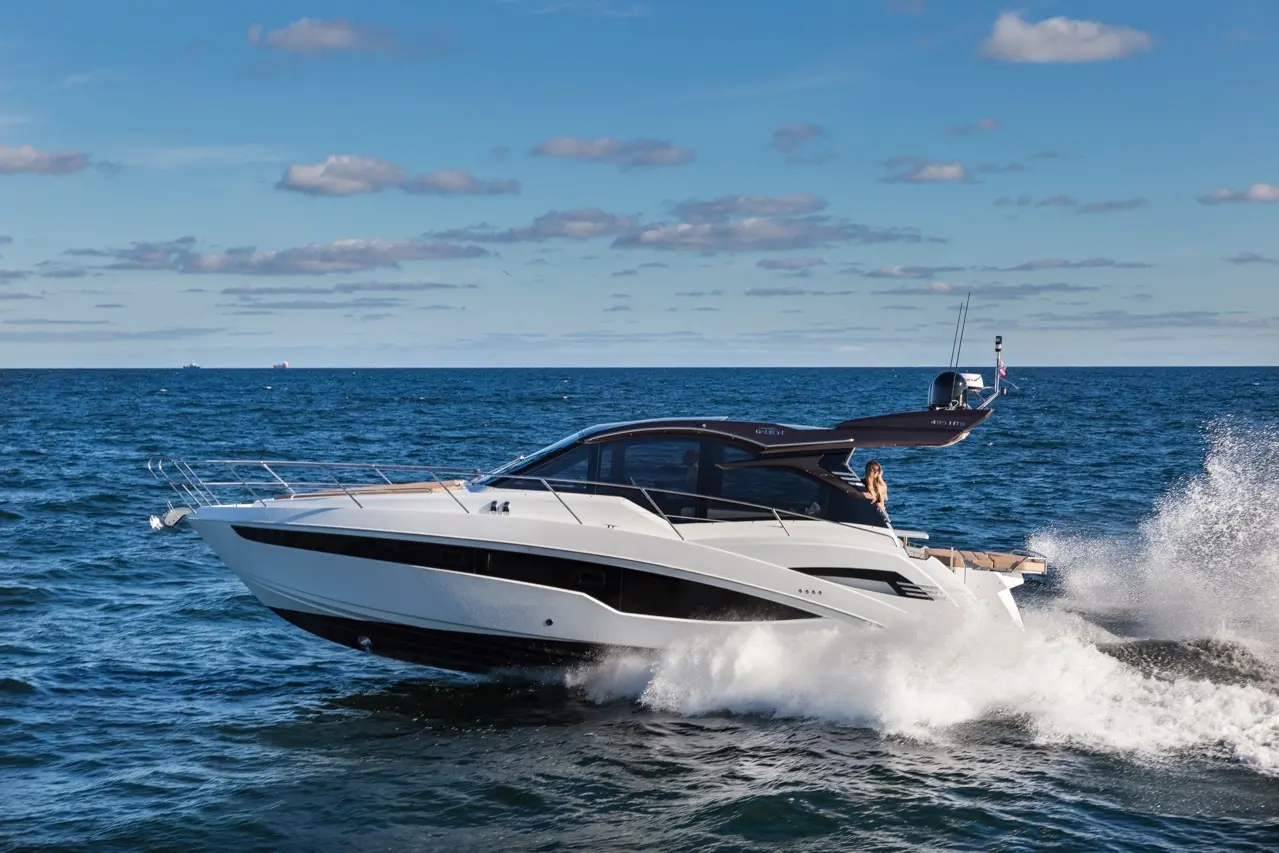 A sleek white luxury yacht speeds across a blue ocean, creating waves; a woman is enjoying the view from the deck.