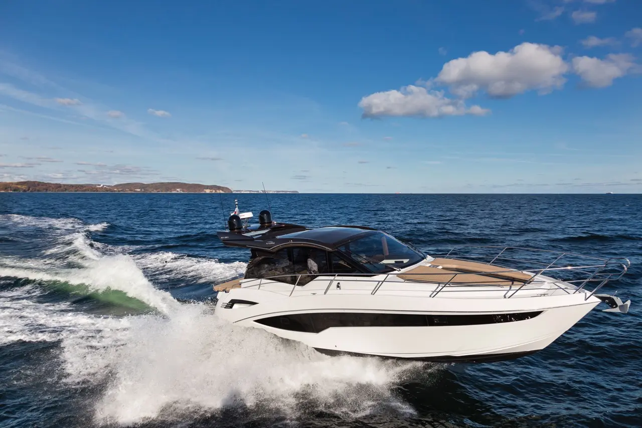 Luxury motorboat navigating through blue waters, creating white spray against a clear sky with distant hills in the background.