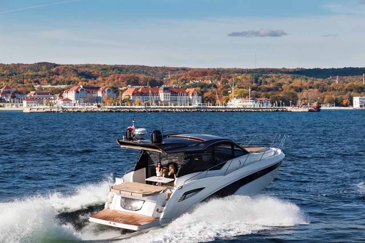 Luxury motorboat with two women onboard speeding through blue water, autumn foliage in the background and a coastal town.