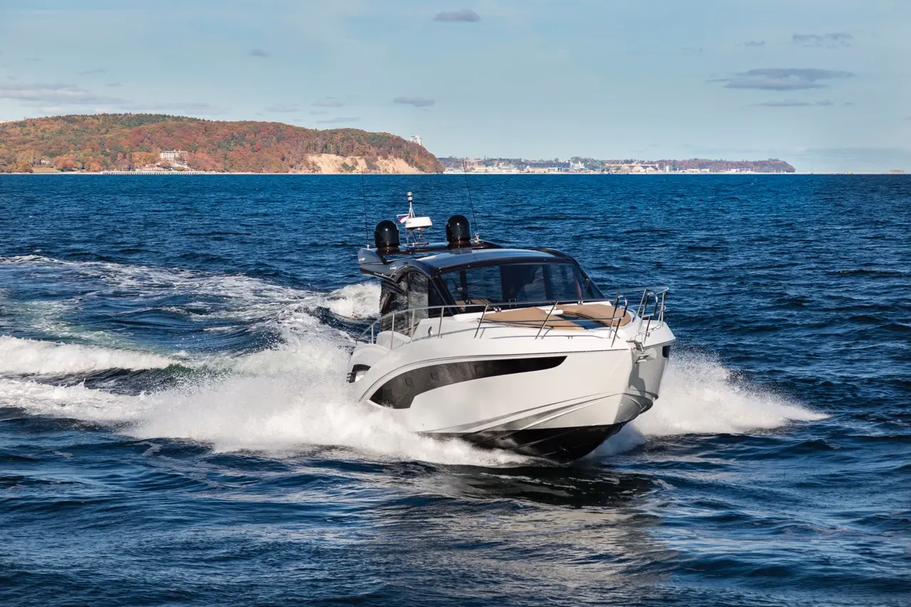 A white motorboat speeds through blue water, leaving foamy trails, with a colorful shoreline visible in the background.