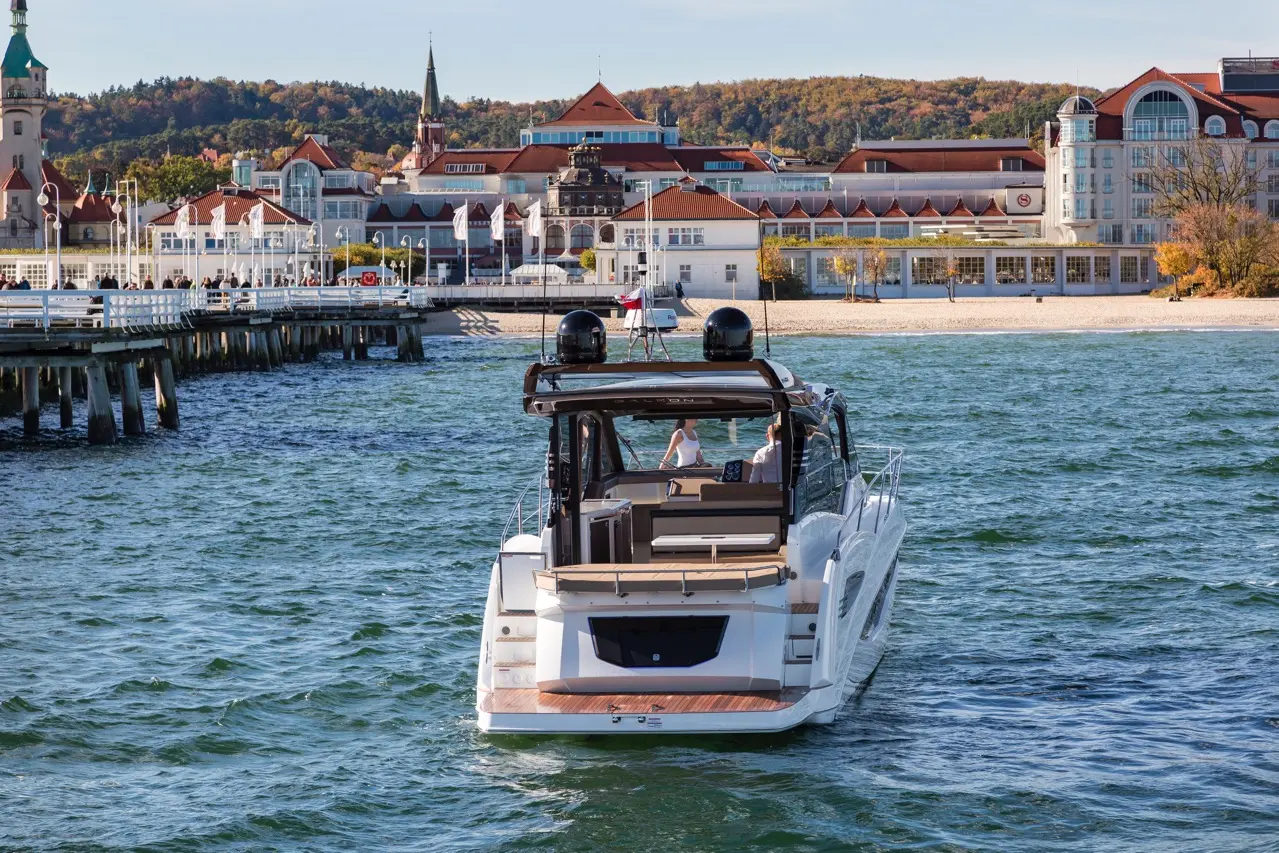 A stylish motorboat navigates clear waters near a scenic waterfront with hotels and autumn foliage in the background.