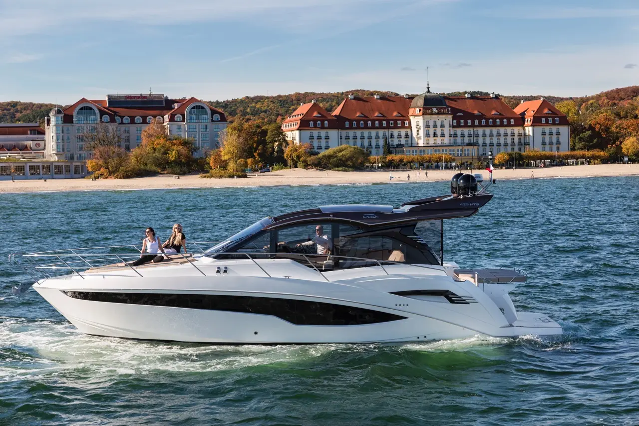 A sleek white motor yacht with passengers sails on water, with a sandy beach and a luxury hotel in the background.