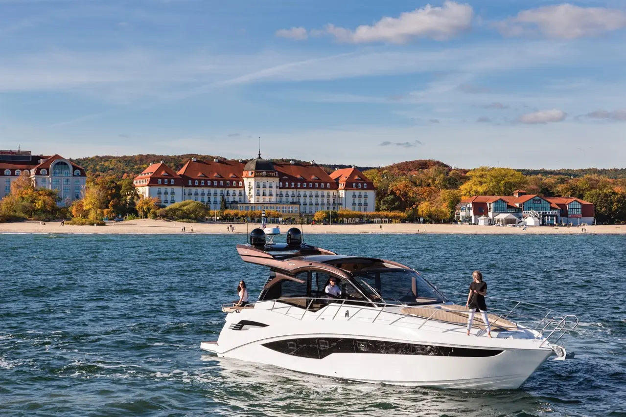 A luxury yacht glides on water near a beachfront hotel, with a woman standing at the bow, surrounded by autumn scenery.